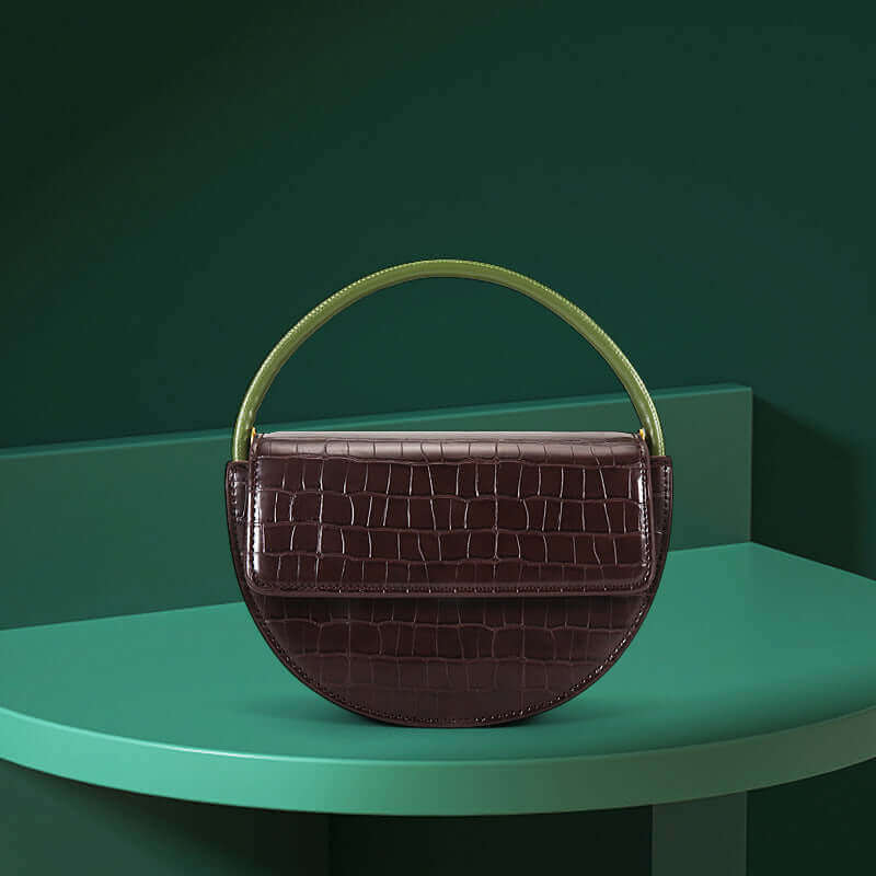 A brown, semi-circle shaped leather handbag with a vibrant green handle, placed on a green surface with a dark background.