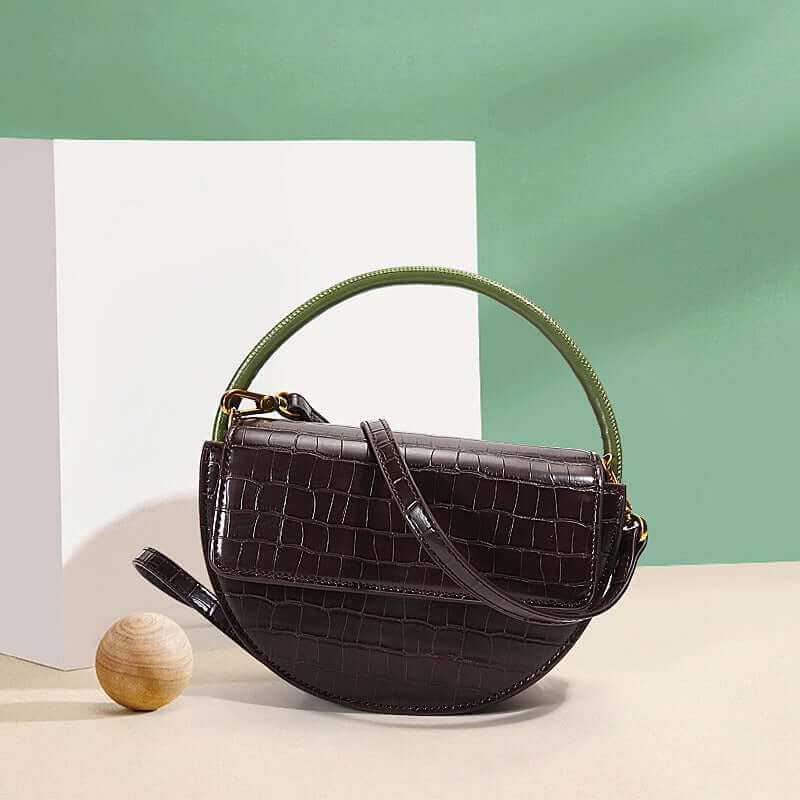 A brown, semi-circle shaped leather handbag with a vibrant green handle, placed on a green surface with a dark background Front Facing