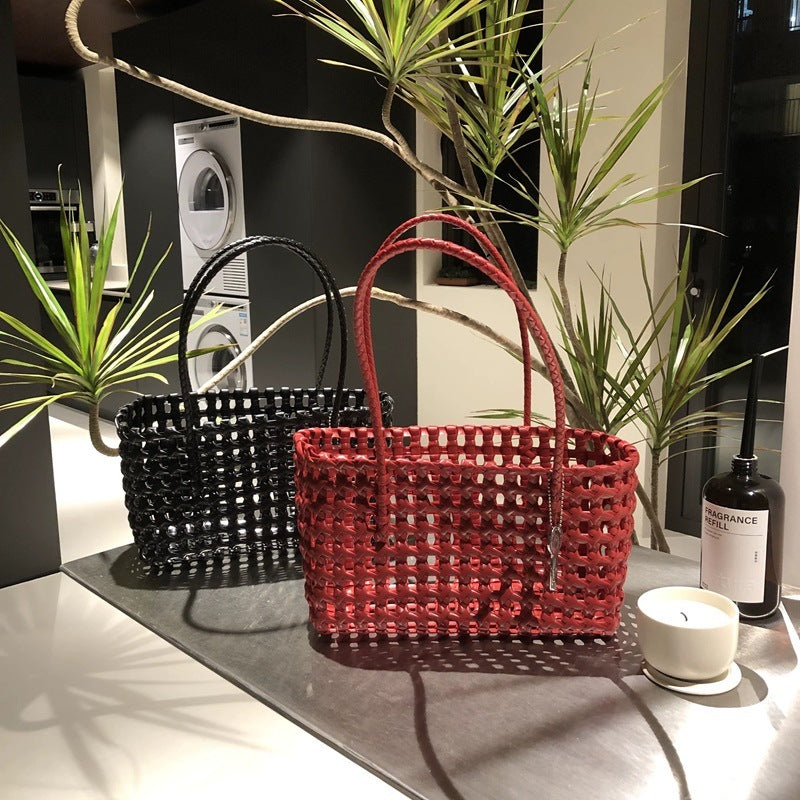 Two woven handbags in red and black colors are displayed on a table with a plant and a bottle, with one bag facing the camera and the other facing away.