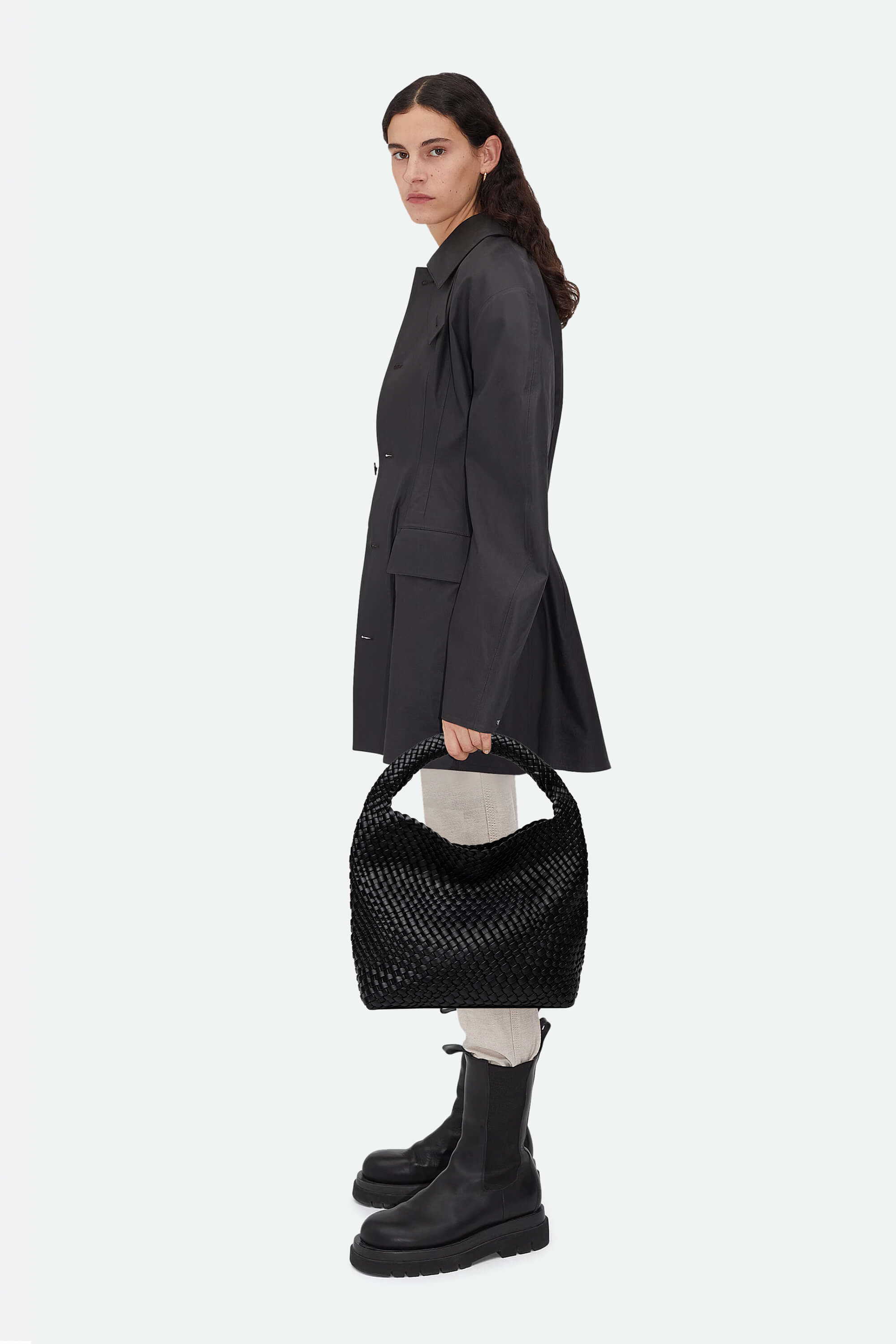 Woman holding a black textured handbag wearing a dark coat and black boots on a white background