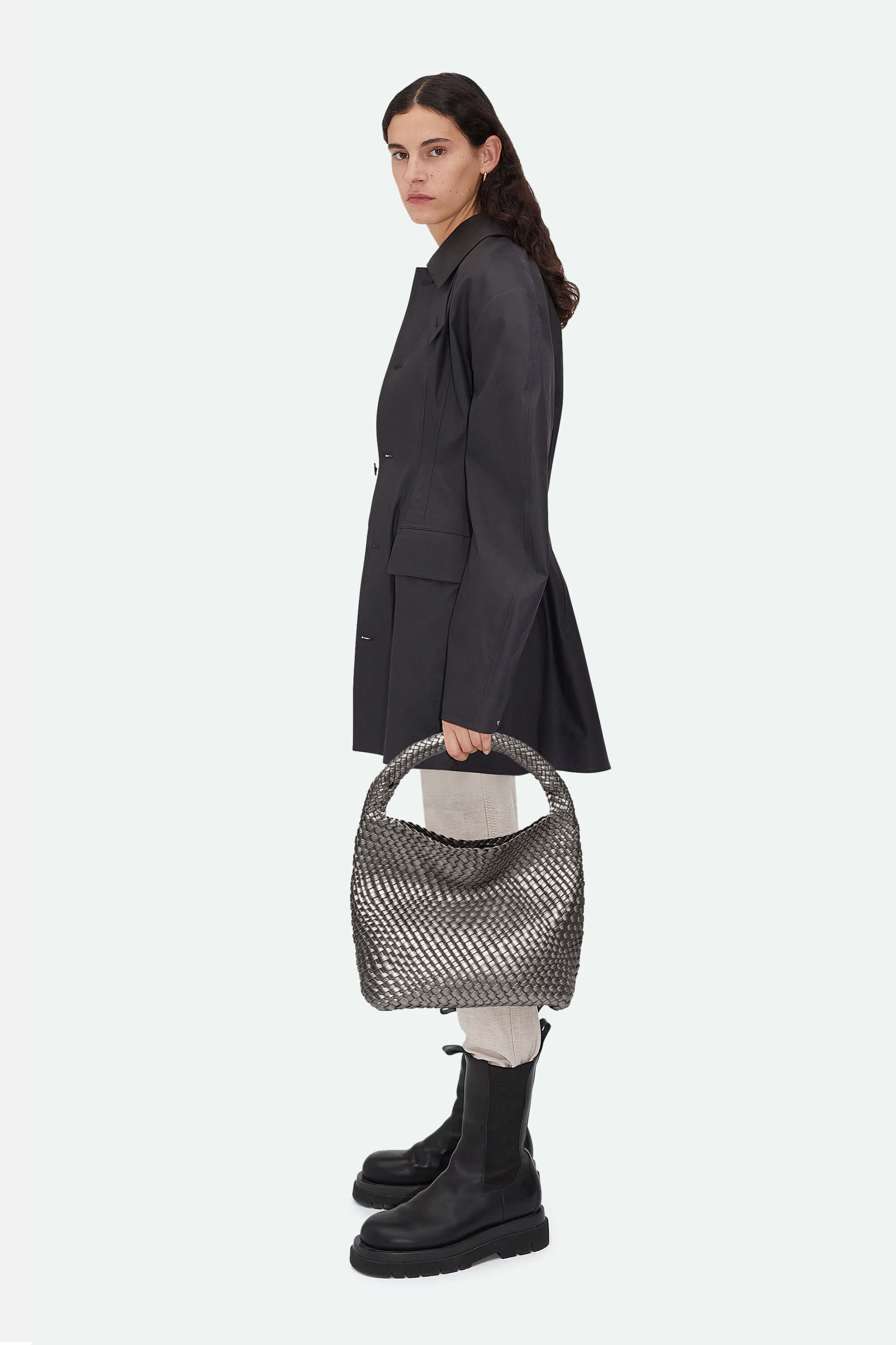 Woman holding a textured handbag, wearing a dark coat and black boots on a white background