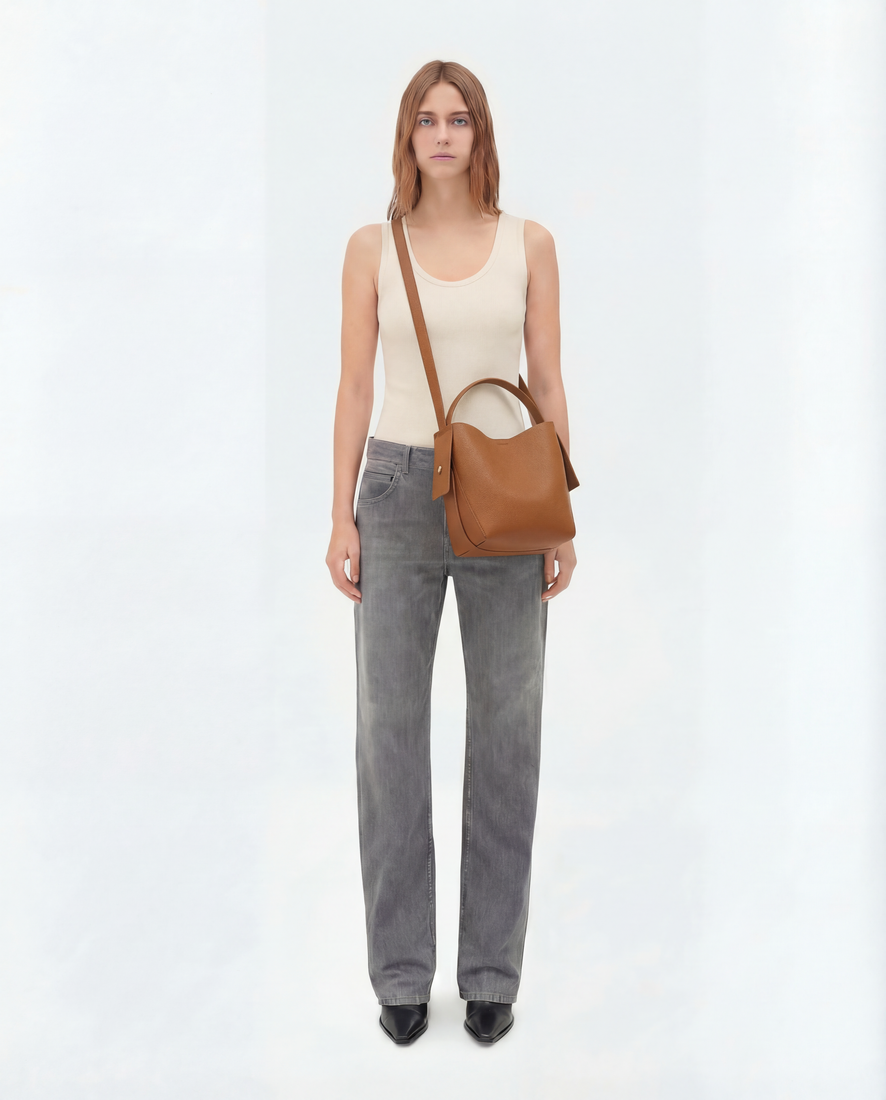 Woman wearing a beige sleeveless top and gray pants, carrying a brown leather bag on a white background