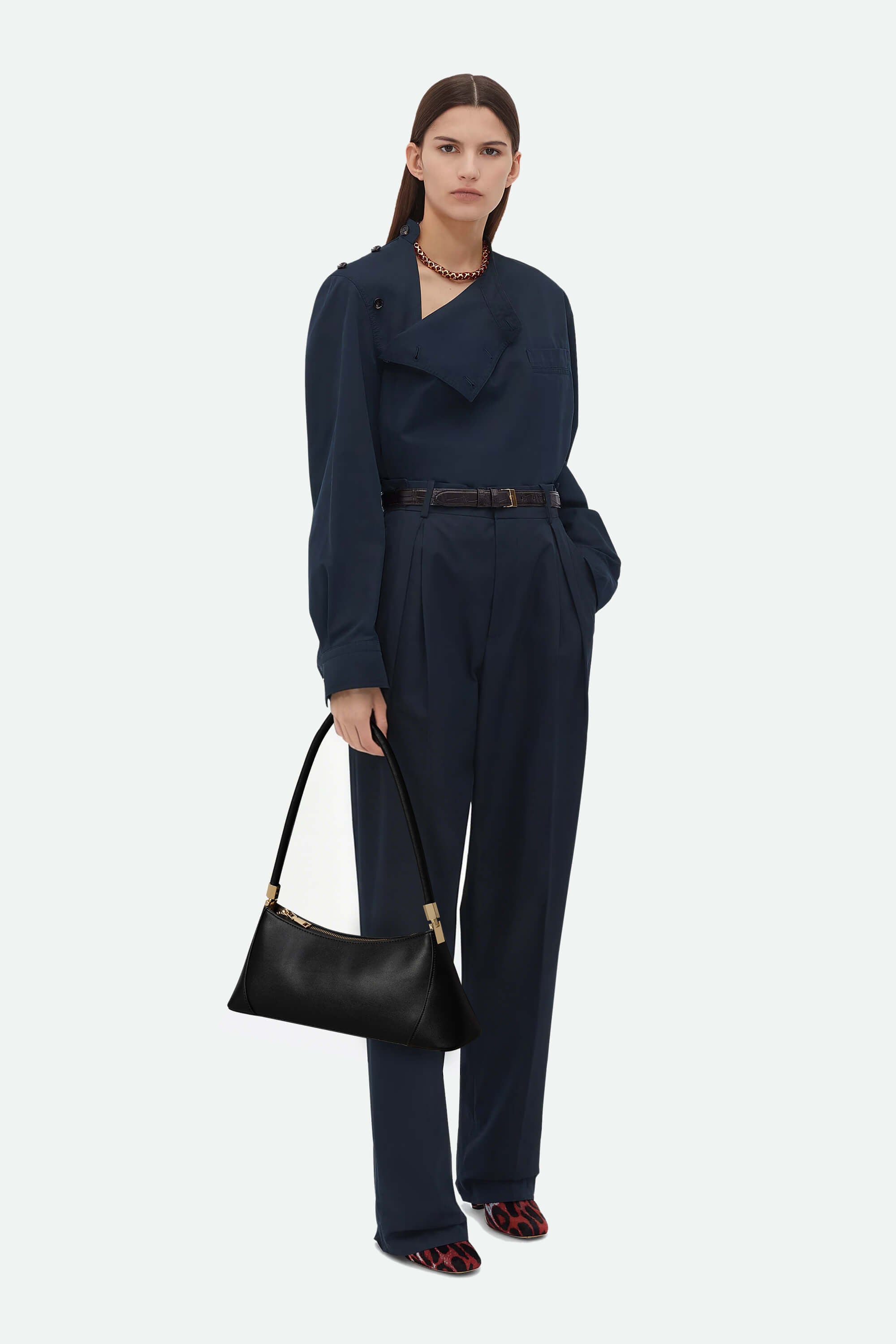 Woman wearing a navy blue jumpsuit holding a black handbag on a white background