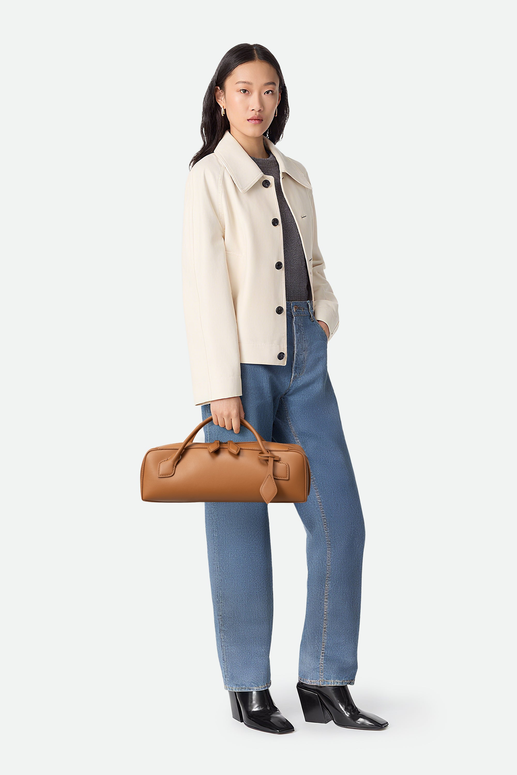 Woman holding a brown leather bag wearing a beige jacket, gray shirt, blue jeans, and black shoes on a white background