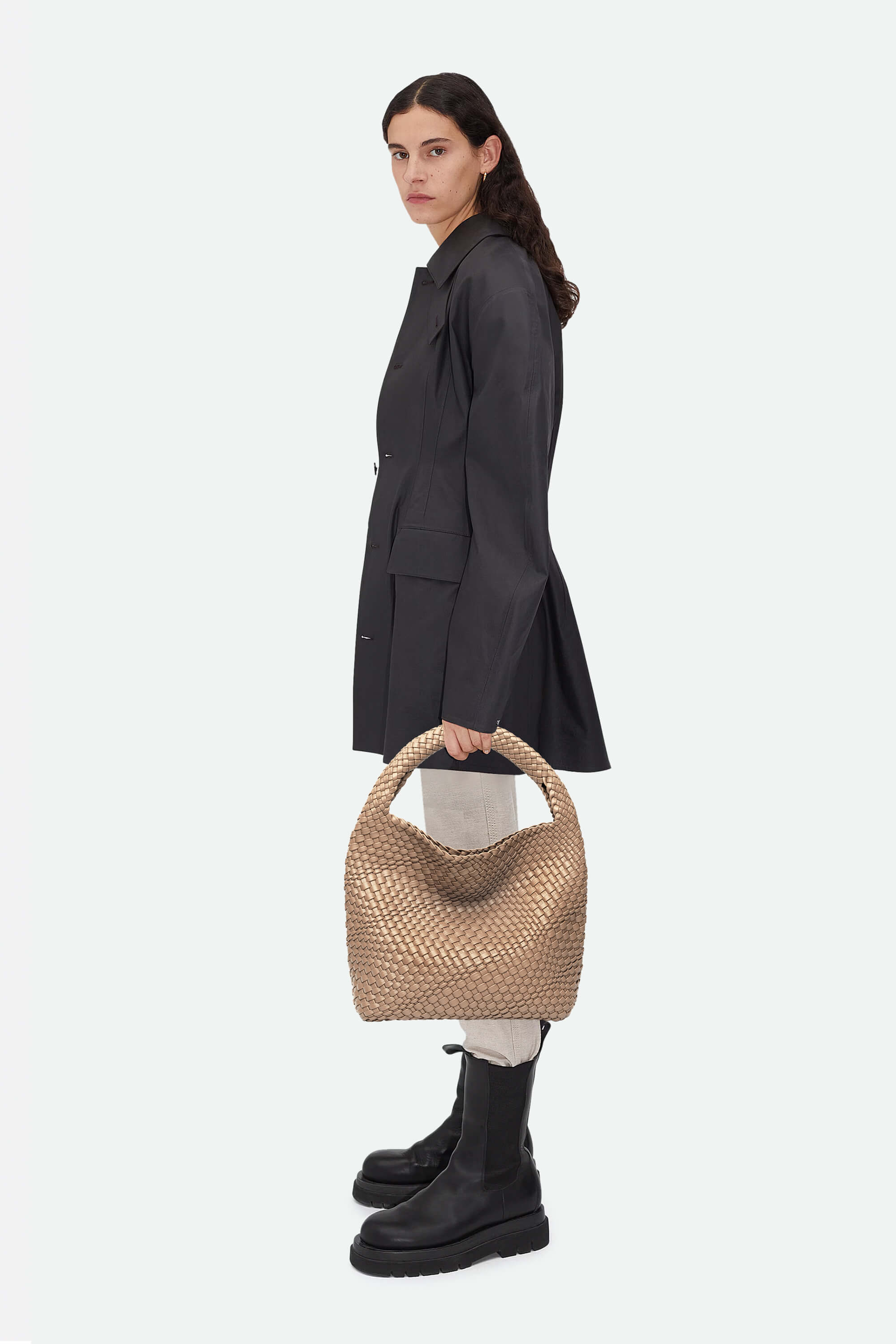 Woman holding a woven handbag wearing a dark coat and black boots on a white background