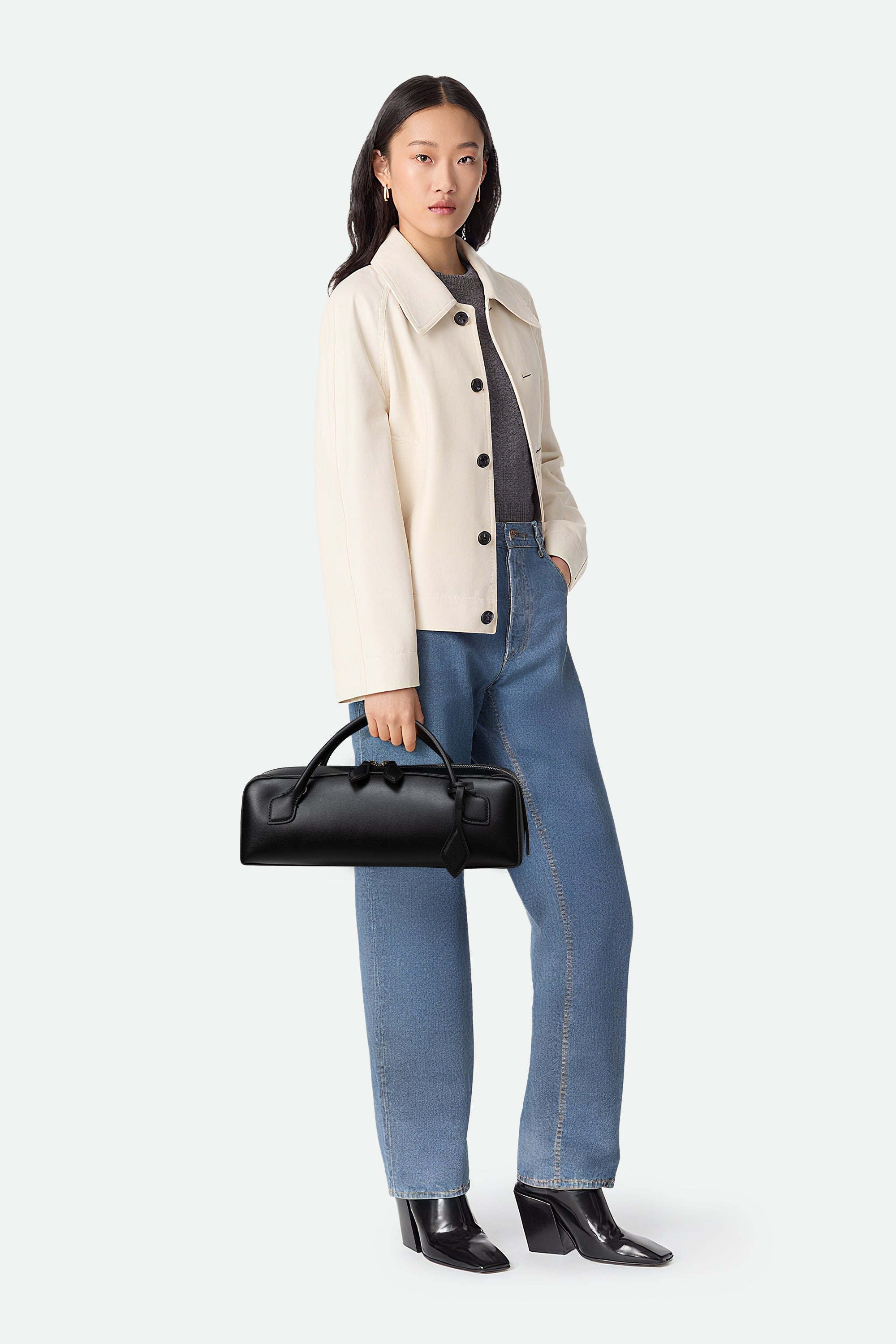 Woman wearing a beige coat, gray shirt, blue jeans, and black shoes, holding a black handbag on a white background.