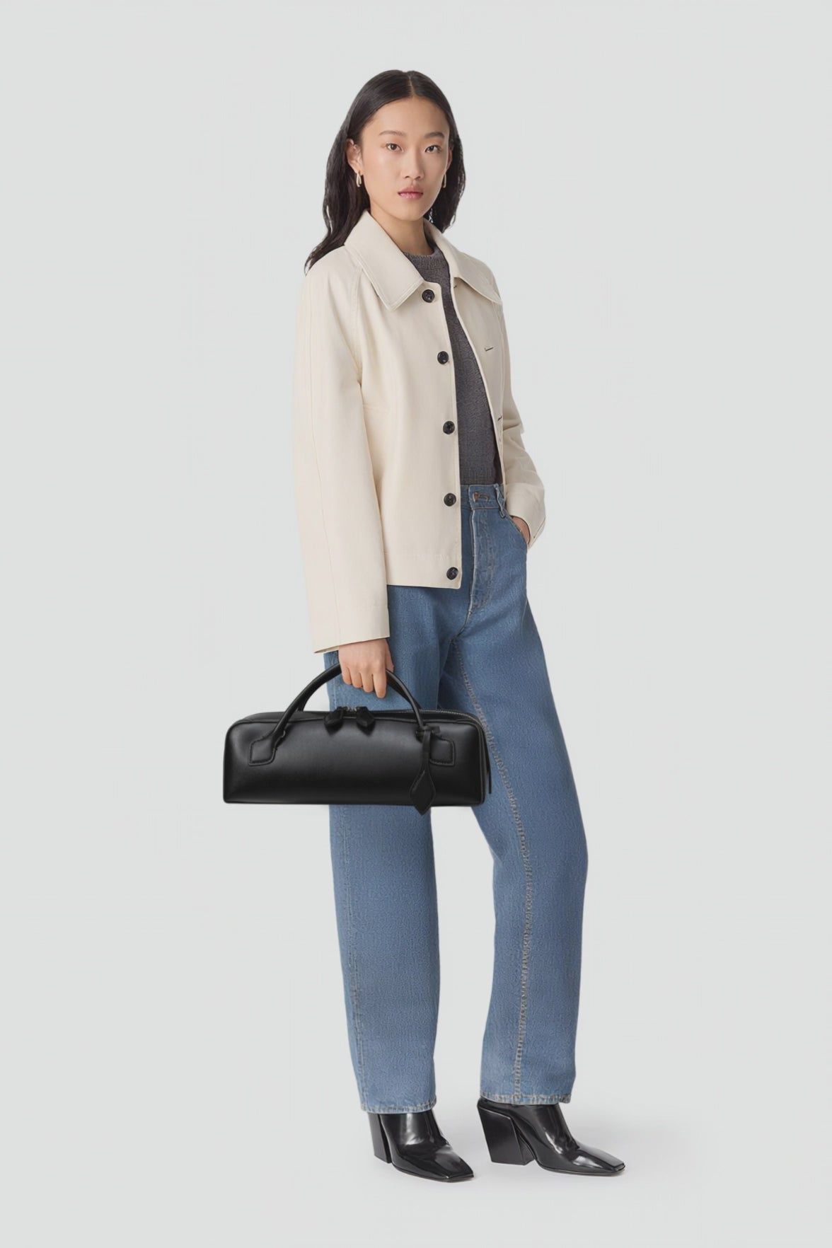Woman wearing a beige coat, gray shirt, blue jeans, and black shoes, holding a black handbag on a white background.