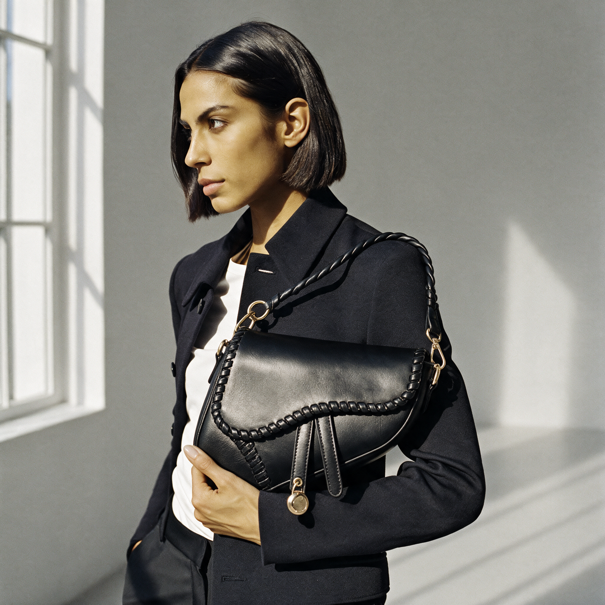 Model in a black jacket carrying a black leather Valentina shoulder bag in a sunlit studio