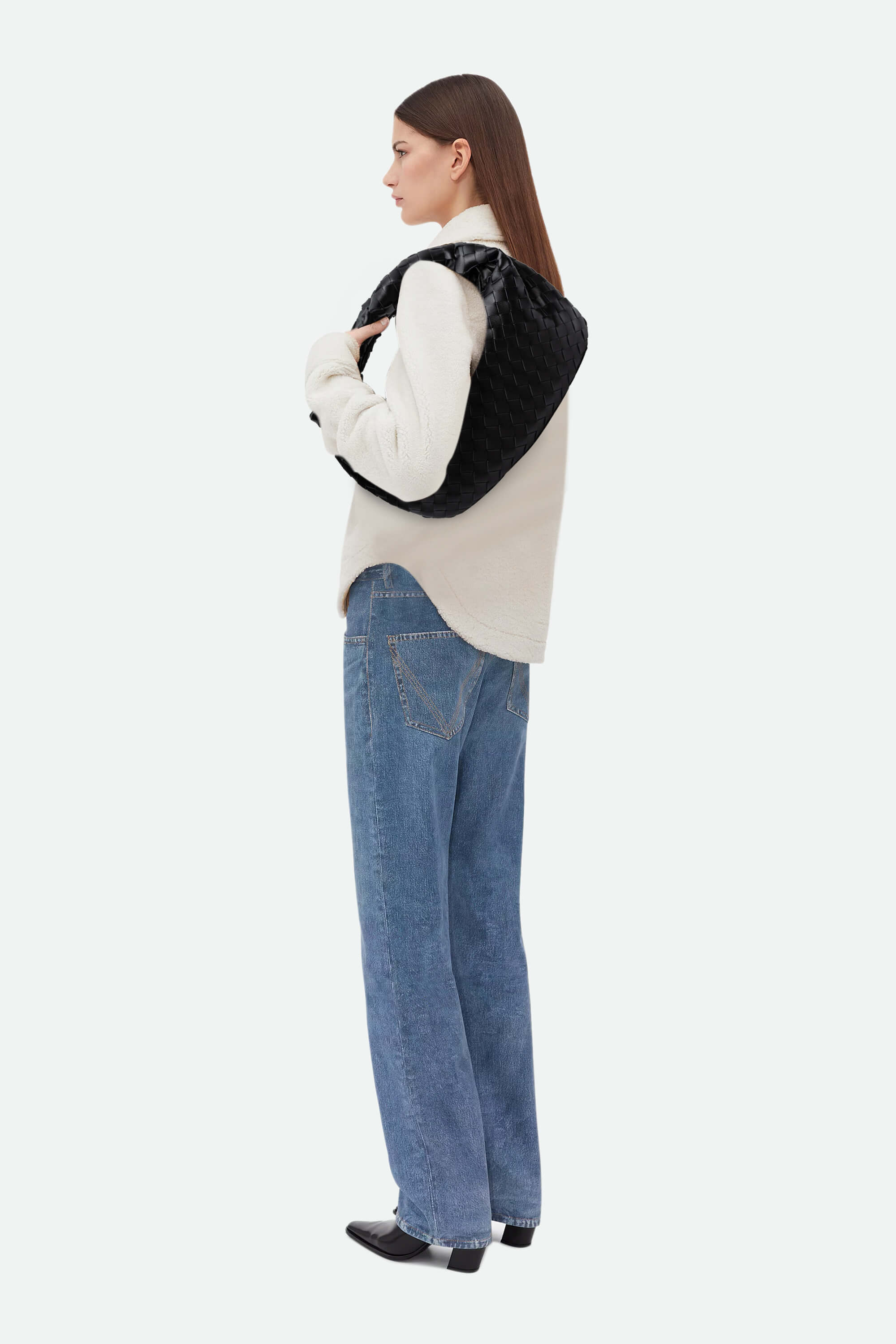 Woman wearing a cream sweater and blue jeans carrying a black woven knot bag on a white background